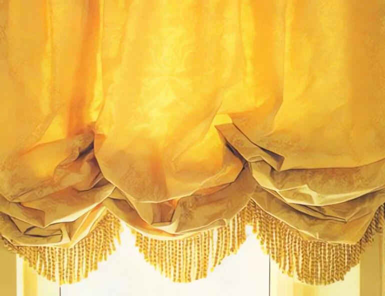 Золотистые шелковые шторы из дамаста собраны в мягкие оборки, кромка подчеркнута золотистой бахромой.